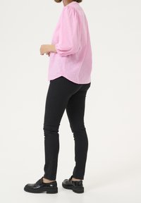 Blouse rose à manches bouffantes avec un motif à pois texturé, associée à un jean noir moulant et des mocassins noirs, vue de côté.