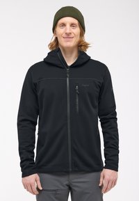 Giacca softshell nera con cappuccio, dotata di chiusura frontale con zip, tasca sul petto con zip e tasche laterali. Texture liscia e design aderente.