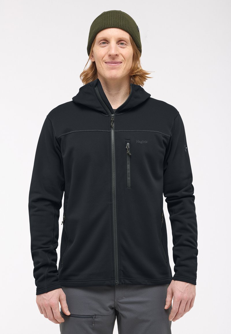 Giacca softshell nera con cappuccio, dotata di chiusura frontale con zip, tasca sul petto con zip e tasche laterali. Texture liscia e design aderente.