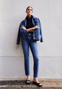 Denim jack en high-waisted jeans in lichtblauw, gecombineerd met zwarte sandalen. Smooth texturen, relaxte pasvorm, minimale details op hardware.