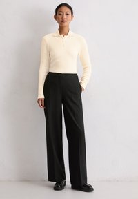Cremfarbener, gerippter Longsleeve-Polooberteil mit Drei-Knopf-Leiste, kombiniert mit hochgeschnittener, schwarzer, weiter Hose und schwarzen flachen Schuhen.