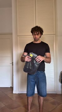 Camiseta preta, calções de ganga azuis, segurando um saco de plástico preto com roupa dobrada dentro, etiqueta amarela visível, em ambiente interno com portas de persiana.