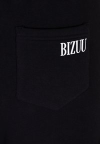 Zwarte stof met een gestructureerde afwerking, voorzien van een vierkante zak en witte "BIZUU" letters boven de zak.