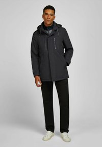 Veste noire avec capuche, deux poches zippées sur la poitrine, fermeture à l'avant et cordons de serrage ajustables. Associée à un pantalon noir et des baskets blanches.