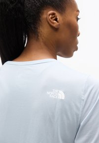 Γυναίκα με σκούρα μαλλιά δεμένα πίσω φοράει ανοιχτό μπλε πουκάμισο με υφή και το λογότυπο The North Face στο πάνω μέρος της πλάτης, κοιτάζοντας προς το πλάι.