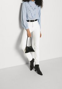 Blusa azzurro chiaro con scollatura a balze, pantaloni bianchi aderenti, stivali neri con punta a strapiombo e una piccola borsa nera.