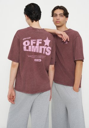Zwei Personen tragen dunkelrote "OFF LIMITS" T-Shirts und hellgraue Hosen, stehen dicht beieinander, eine Person hat den Arm auf der Schulter der anderen.