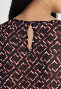 Blouse noire à motif présentant un design géométrique rouge et blanc, avec une ouverture en forme de clé à l'arrière et une texture de tissu léger.