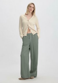 Beige geribde cardigan met v-hals en knoopsluiting, gecombineerd met losse groene broeken. Zachte textuur en neutrale kleuren creëren een ontspannen uitstraling.