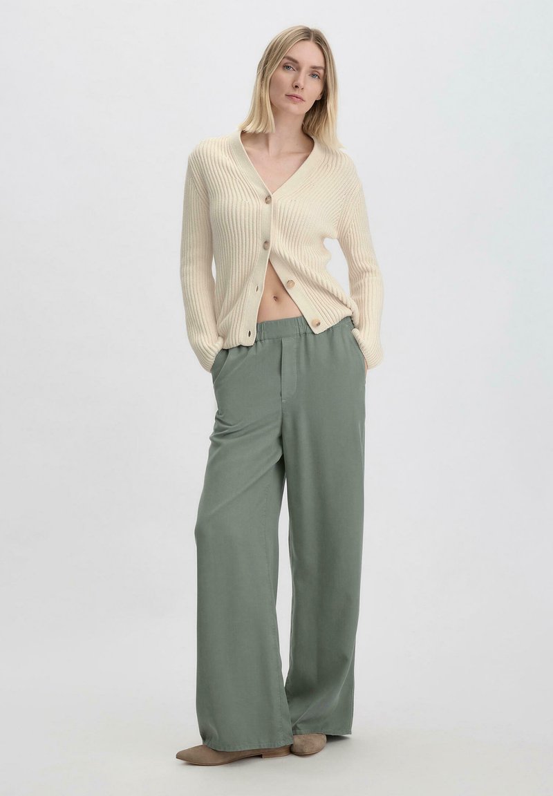 Beige geribde cardigan met v-hals en knoopsluiting, gecombineerd met losse groene broeken. Zachte textuur en neutrale kleuren creëren een ontspannen uitstraling.