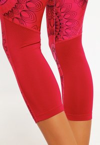 Tajta byxor i livlig röd med mönstrade sektioner som har svarta blommönster och texturerade nätinsatser på de nedre panelerna. Elastisk midja.