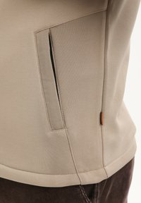 Primer plano del bolsillo de una chaqueta beige con un forro interior negro visible y una pequeña etiqueta naranja, llevada sobre pantalones de ante marrón oscuro.