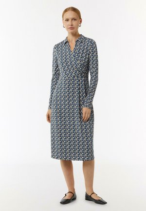 Langarm wikkeljurk in navy met een geometrisch beige patroon, voorzien van een kraaghals en een getailleerde pasvorm, gecombineerd met zwarte platte schoenen.