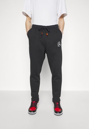 Person, der schwarze Jogginghosen mit Kordelzug und Logo am Oberschenkel, ein weißes T-Shirt und schwarz-rote Sneaker trägt, steht vor weißem Hintergrund.