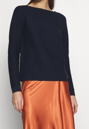 Kvinde iført en marineblå langærmet sweater kombineret med et skinnende orange satin-nederdel, stående foran en ensartet lys baggrund.