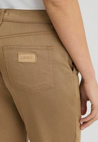 Gros plan sur une personne portant un pantalon marron avec une poche arrière étiquetée "LIU JO" et sa main droite reposant à côté de sa hanche.