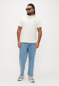 Lysebeige t-shirt med lille sort logo på venstre bryst, kombineret med blå løstsiddende jeans og grå sneakers. Solbriller fuldender looket.