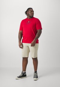 T-shirt rossa a maniche corte con un disegno grafico sul petto, abbinata a pantaloni cargo beige e sneaker alte nere.