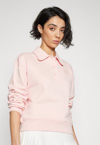 Sudadera de polo rosa claro con cuello tipo polo, tapeta de tres botones y puños acanalados. Presenta un sutil detalle del logo en el pecho.