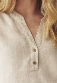 Béžová lněná košile s výstřihem do V a dvěma dřevěnými knoflíky, kterou nosí osoba s dlouhými blond vlasy.