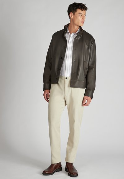 Giacca di pelle marrone, abbottonata, con una camicia a righe verticali, abbinata a pantaloni chiari e stivali alla caviglia marroni.