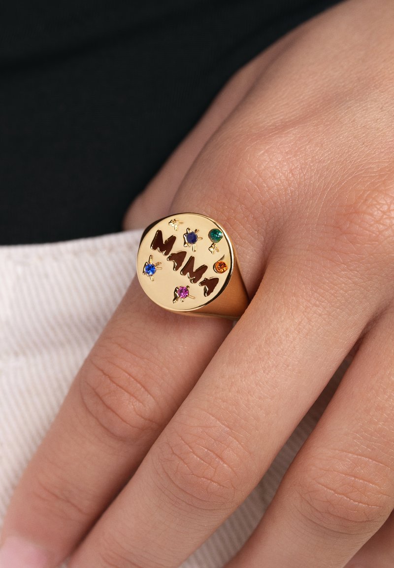 Anillo en tono dorado con la palabra "Mamá" en letras recortadas, adornado con coloridas piedras preciosas en azul, verde, púrpura, naranja y rosa.