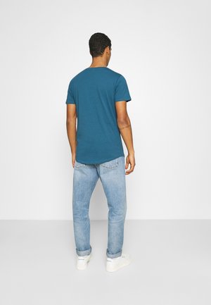 T-Shirts in Türkis mit kurzen Ärmeln und Rundhalsausschnitt, kombiniert mit hellblauen Jeans und weißen Sneakern, vor einem einfarbigen Hintergrund präsentiert.