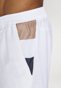 Shorts blancs avec une poche latérale présentant un design en maille superposée beige, blanche et bleu marine. Ceinture élastique avec des détails texturés.