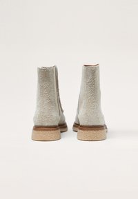 Bottines grises texturées avec une finition lisse, semelle plate en caoutchouc marron clair, et panneaux élastiques latéraux pour un enfilage facile. Vue de dos affichée.