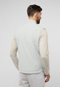 Gilet grigio chiaro con una texture liscia e leggera, chiusura frontale con zip e senza maniche, indossato sopra una camicia beige a maniche lunghe.