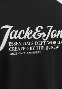 Camiseta de algodón negra con texto blanco. El diseño incluye "Jack & Jones" en una fuente cursiva y detalles adicionales en una fuente sans-serif.