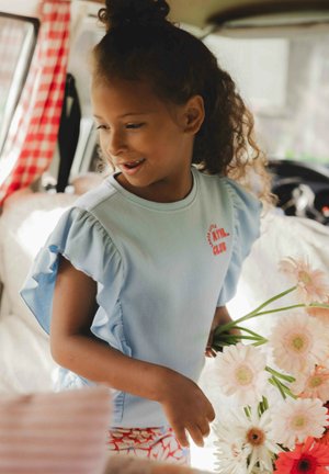 Lichtblauw geribd T-shirt met geschulpte korte mouwen en een rode grafische tekst. Gecombineerd met rode bloemenpatroon shorts, terwijl ze witte en roze bloemen vasthoudt.