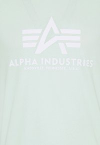 Gaiši zaļš T-kreklis, izgatavots no mīksta auduma, ar izteiktu baltu logotipu, uz kura ir rakstīts "ALPHA INDUSTRIES" un zem tā ir norādītas atrašanās vietas detaļas.