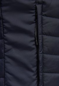 Mörkblå puffjackematerial med vadderad struktur. Har en svart dragkedja med ett dragskydd och linjära sydda detaljer längs sömmarna.