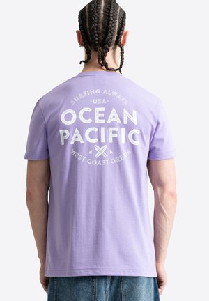 Uomo con capelli intrecciati che indossa una t-shirt lavanda con testo bianco "Ocean Pacific" e grafica di tavola da surf sul retro, abbinata a jeans blu.