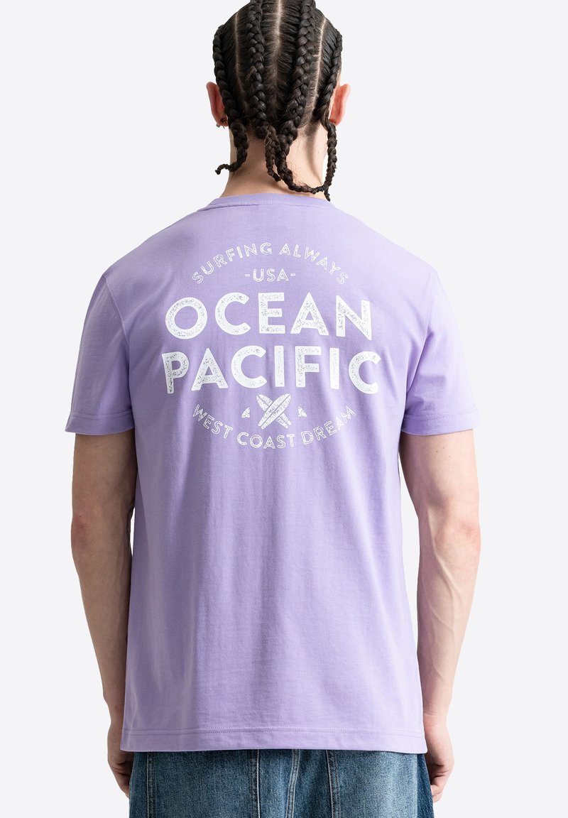 Vyras su pintais plaukais dėvintis levandų spalvos marškinėlį su baltais žodžiais „Ocean Pacific“ ir banglentės grafika ant nugaros, derinamas su mėlynomis džinsomis.