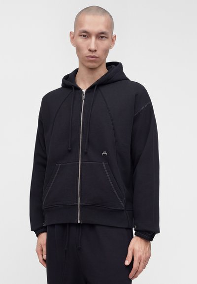 Άνδρας που φοράει μαύρο φούτερ με φερμουάρ, εμπρός τσέπες και κορδόνια, στέκεται μπροστά σε απλό ανοιχτόχρωμο φόντο.