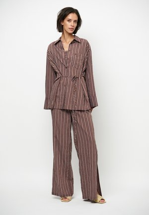 Femme debout portant une chemise ample à rayures verticales brunes et blanches avec un pantalon large assorti et des talons beiges à bout ouvert sur fond blanc.