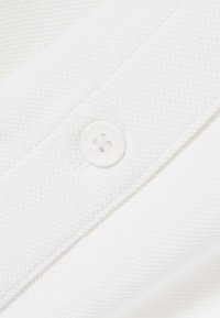 Nahaufnahme eines weißen, strukturierten Stoffes mit einem angenähten, runden weißen Knopf mit vier Löchern.