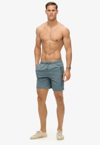 Heren zwembroek in teal stof met een matte afwerking, voorzien van een elastische tailleband met trekkoord en het "Superdry" logo aan de zijkant.
