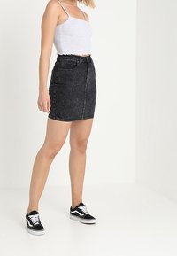 Falda de mezclilla negra de cintura alta con un lavado desgastado, bolsillos delanteros y corte recto, combinada con una camiseta sin mangas corta de color gris claro y zapatillas negras.