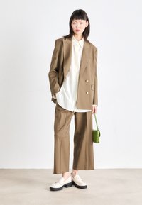 Bruin maatpak met een dubbelzijdige blazer en wijde pijpen. Witte blouse eronder, gecombineerd met crèmekleurige schoenen en een groene handtas.