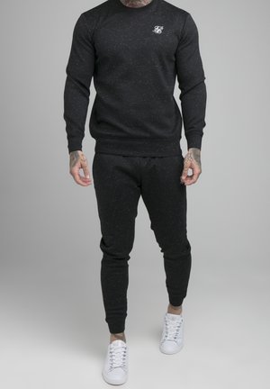 Mann trägt schwarzen, gesprenkelten Pullover und passende Jogginghosen mit weißen Sneakern, steht vor einem schlichten grauen Hintergrund.