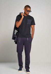 Zwart t-shirt met een blauwe zak, gecombineerd met donkerpaarse joggingbroek met reflecterende accenten, en blauwe instap schoenen.