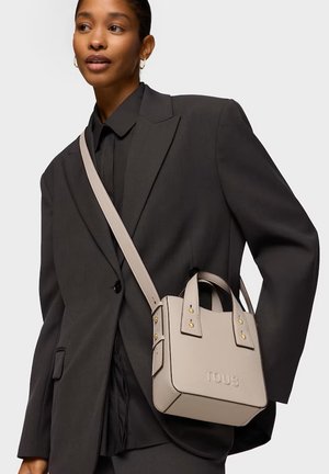 Mujer con traje negro y bolso TOUS beige llevado cruzado sobre su hombro frente a un fondo liso.