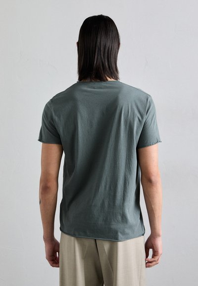 Green cotton t-shirt with short sleeves, relaxed fit, crew neckline, and a slightly curved hem, paired with light beige trousers.