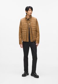 Brūns kviltots mētelis ar augstu apkakli, melnām pogām un gludu apdari, apvienots ar melniem biksēm un melniem šņorējamiem apaviem.