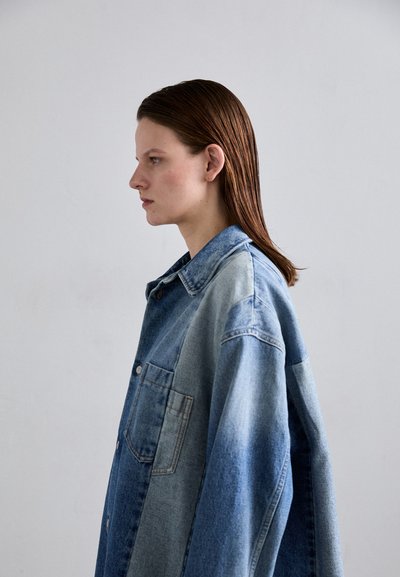 Gaiši zila denima oversized krekls ar pogu priekšpusē, ar kabatu un dažādām denima toņu un tekstūru kombinācijām.