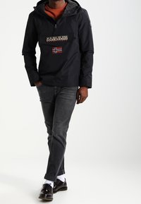 Czarna wodoodporna kurtka z kapturem, wyposażona w przednią kieszeń, logo oraz detal z norweską flagą, w połączeniu ze szarymi dżinsami i ciemnymi butami.