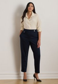 Krémová blúzka s golierom, priliehavé tmavomodré nohavice s vysokým pásom a s gombíkovými detailami, doplnené čiernymi podpätkami. Hladká textúra látky.